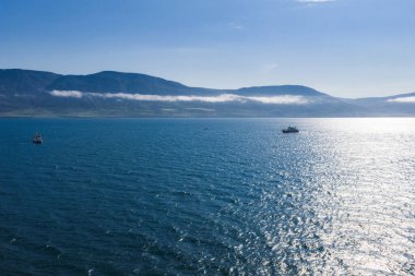Mavi bir okyanusun sakin hava manzarası. Arka planda alçak uzanan dağlar, sisli bulutlar ve suya yansıyan güneş ışığı..
