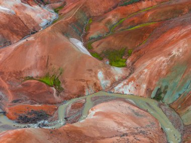 İzlanda 'nın Gökkuşağı Dağları' nın canlı kırmızı, turuncu ve yeşil renkleri, dolambaçlı bir akarsu, yeşil yosun ve beyaz mineral yataklarının yer aldığı hava manzarası..
