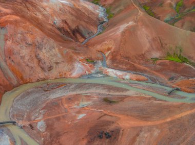 İzlanda 'nın Gökkuşağı Dağları' nın canlı kırmızı, turuncu ve kahverengi renkleriyle hava manzarası. Sarmal bir nehir ve ahşap köprüler engebeli arazide ilerliyor..