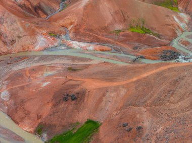 Sarmal yolları olan canlı rhyolite tepeleri, derenin üzerinde ahşap bir köprü ve kırmızımsı turuncu ve bej arazi ile çelişen yeşil bitki örtüsü..