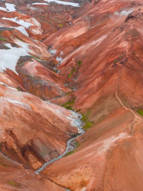 Landmannalaugar, İzlanda 'daki parlak kırmızı, turuncu ve kahverengi rhyolite dağları. Kar yamaçları, buhar bacaları, dolambaçlı bir patika ve küçük bir deresi var..