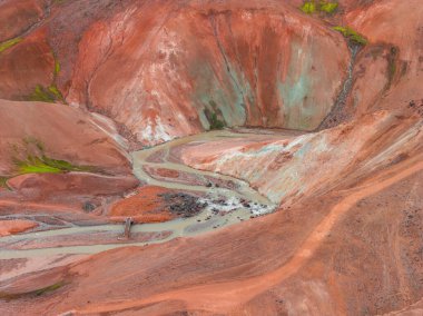 İzlanda 'nın Gökkuşağı Dağları' nın hava manzarasında canlı kırmızı, turuncu ve yeşil renkler, dolambaçlı bir dere ve onu geçen küçük ahşap bir köprü var..