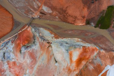 Turuncu, kırmızı ve beyaz mineral yatakları, çamurlu bir nehir, küçük bir köprü ve yeşil bitki örtüsünün yer aldığı İzlanda jeotermal arazisinin hava görüntüsü..