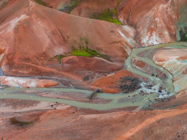 İzlanda 'nın Gökkuşağı Dağları' nın canlı kırmızı, turuncu ve yeşil renkleriyle hava manzarası. Tahta köprüsü ve küçük figürleri olan bir nehir görülebilir..