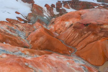 İzlanda 'nın Gökkuşağı Dağları' nın hava manzarasında canlı turuncu ve kırmızı ryolit oluşumlar, kar yamaları ve engebeli arazide dolambaçlı bir yol var..