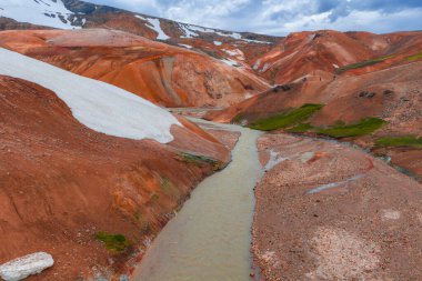 İzlanda 'nın Gökkuşağı Dağları' nın canlı kırmızı, turuncu ve kahverengi renkleri yeşil bitki örtüsü, kar bantları ve renkli araziyi kesen bir nehir..