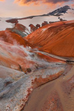 Kerlingarfjoll 'un buhar delikli kırmızı ve turuncu rhyolite dağları, derenin üzerinde ahşap bir köprü, karlı zirveler, ve gün batımında parlayan bir ışık..