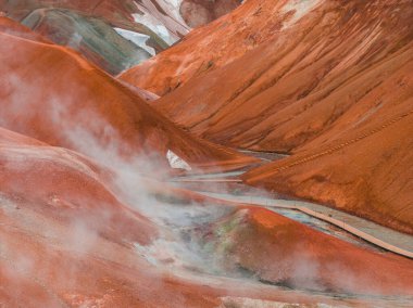 Jeotermal bacalardan yükselen buharlı kırmızı ve turuncu ryolit dağları, bir ahşap patika ve Kerlingarfjoll, İzlanda 'da kar yamaları.