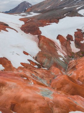 İzlanda 'nın Gökkuşağı Dağları' nın canlı kırmızı, turuncu ve kahverengi ryolit oluşumları, arka planda engebeli bir tepe ile beyaz karla tezat oluşturur..