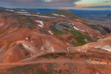 İzlanda 'nın Gökkuşağı Dağları' nın canlı kırmızı, turuncu ve yeşil renkleri, dağınık kar parçacıkları ve ön planda bir park alanı yer alıyor..