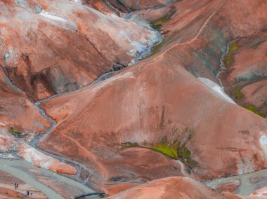 Kırmızı, turuncu ve kahverengi tonlarında canlı rhyolite tepeleri, yeşil yosun ve kar yamaçları. Küçük rakamlar manzaranın ölçeğini vurgular.