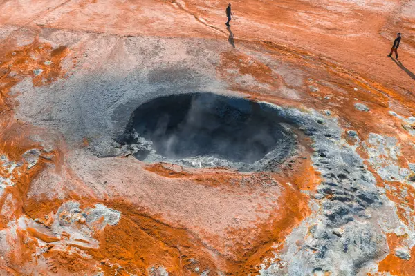 İzlanda 'da buharlaşan bir jeotermal kaplıcanın havadan görünüşü, turuncu ve beyaz mineral yataklarıyla çevrili. Yakınlardaki kırmızı arazide iki kişi duruyor..