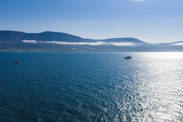 Mavi bir okyanusun sakin hava manzarası. Arka planda alçak uzanan dağlar, sisli bulutlar ve suya yansıyan güneş ışığı..
