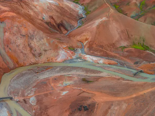 İzlanda 'nın Gökkuşağı Dağları' nın canlı kırmızı, turuncu ve kahverengi renkleriyle hava manzarası. Sarmal bir nehir ve ahşap köprüler engebeli arazide ilerliyor..