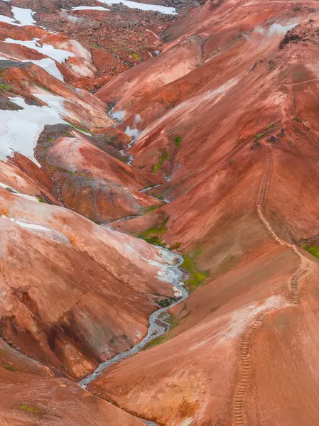 Landmannalaugar, İzlanda 'daki parlak kırmızı, turuncu ve kahverengi rhyolite dağları. Kar yamaçları, buhar bacaları, dolambaçlı bir patika ve küçük bir deresi var..