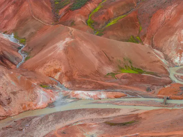İzlanda 'nın Gökkuşağı Dağları' nın çok renkli ryolit tepeleri. Kırmızı, turuncu ve yeşil renkleri var. Ahşap bir köprü nehri geçiyor, yakında buhar yükseliyor..