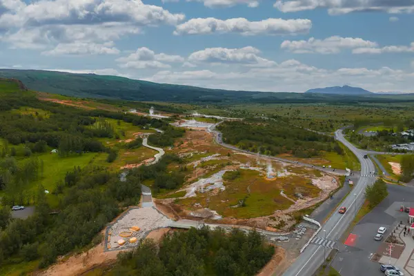 İzlanda 'daki Geysir Jeotermal Bölgesi' nin hava manzarası, dumanı tüten bacalar, dolambaçlı yollar, yemyeşil yollar ve bulutlu bir gökyüzünün altındaki uzak dağlar..