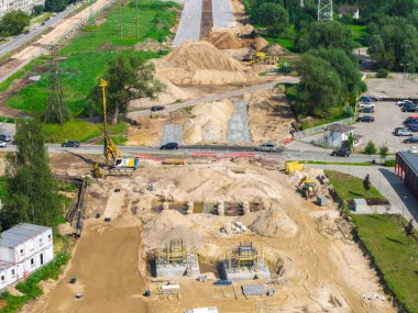 Riga 'dan Jelgava yoluna kadar büyük ölçekli yol inşaatı sondaj kulesi, kum yığınları ve kısmen asfalt bir yol. Yeşil alanlar, araçlar ve geçici binalar görünür.