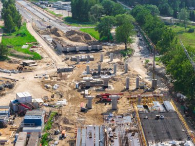 Riga 'dan Jelgava yoluna kadar büyük ölçekli inşaat alanı beton sütunlar, ağır makineler ve malzemelerle. Etrafı yeşilliklerle çevrili, kısmen inşa edilmiş bir yol görünür..