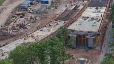 Hareket halindeki araçlar, işçiler ve ağır ekipmanlarla büyük ölçekli yol inşaatı. Kırmızı bir bina, iletişim kulesi ve yeşillik görünüyor..