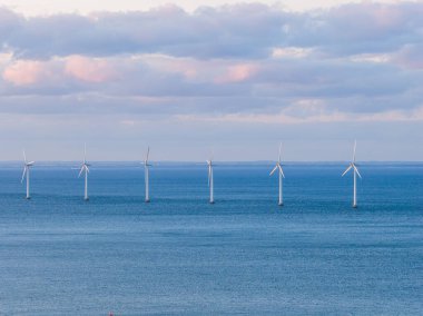 Danimarka, Kopenhag 'da sürdürülebilir enerji altyapısını gösteren, pembe renkli, parçalı bulutlu bir gökyüzünün altında altı rüzgar türbini durgun mavi sularda duruyor..