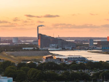 Kopenhag, Danimarka 'nın hava manzarası, gün batımında kayak pisti çatısı, bacası, su yolu, sanayi binaları ve yeşil alanları ile KopenHill fabrikası.