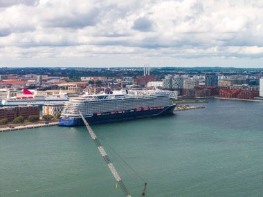 Kopenhag limanına demirlemiş bir yolcu gemisinin hava görüntüsü, modern liman binaları, Amager Bakke tesisi, sakin su ve görünür bir vinç..