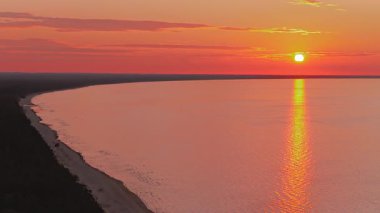 Sakin bir denizin üzerinde sakin bir günbatımı, sudaki canlı turuncu yansımaları ve kıyı şeridi boyunca dokulu bir gökyüzünün altında hareket eden nazik dalgalarla..