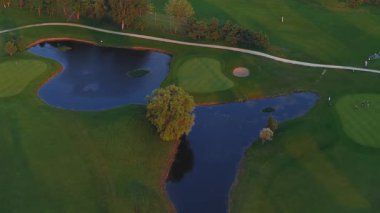 Gün batımında bir golf sahasının hava görüntüsü, yeşil çim yolları, yansıtıcı havuzlar, belirgin bir ağaç ve dolambaçlı yollar..