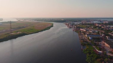 Letonya 'da günbatımında sakin bir nehir manzarası, bir kıyıda endüstriyel yapılar, diğerinde yeşil alanlar, yavaş çekimde yakalandı..