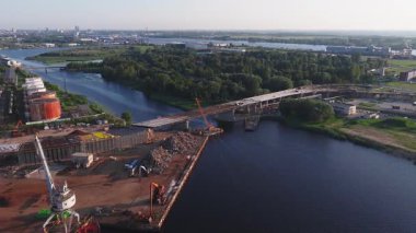 Riga, Letonya 'daki inşaat sahasında, arka planda kentsel ve endüstriyel unsurların yer aldığı, nehir üzerinde köprü inşa eden vinç ve makineler görülüyor..