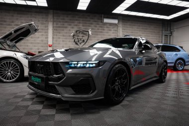 Gri Ford Mustang GT 5.0. Parlak kapalı bir galeride siyah alaşımlı tekerler. Bir Mercedes AMG sınıfı ve Bentley arka planda kısmen görünür..