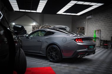 Gri Ford Mustang GT 5.0. Bagajı açık, arka farları açık ve çift egzozlu. İyi aydınlatılmış bir garajda, kareli zemini ve görülebilir aletleri var..
