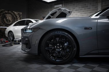 Gri Ford Mustang GT 5.0 siyah alaşımlı tekerlekler ve odaklı Brembo frenleri. Sanayi ortamında açık kapüşonlu beyaz Mercedes AMG sınıfı..