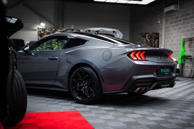 Kırmızı halı aksanlı, kareli zeminde gri bir Ford Mustang GT 5.0, üçlü bar LED stop lambaları, çift egzozlar, ve modern bir sergi salonunda bir spoiler..
