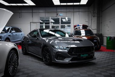 Gri bir Ford Mustang GT 5.0. Galeride keskin LED farları var. Arka planda Mercedes AMG sınıfı, Bentley ve kareli zemin görünüyor..