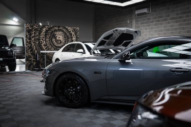 Gri bir Ford Mustang GT 5.0, modern bir garajda kareli zemine park etmiş siyah tekerlekleri var. Siyah bir Mercedes AMG sınıfı ve beyaz bir araba görünüyor..