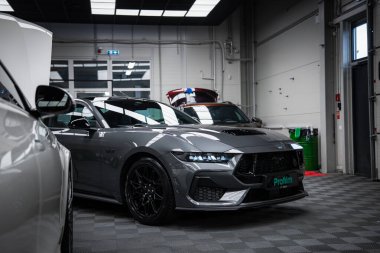 Gri bir Ford Mustang GT 5.0 siyah alaşımlı tekerler ve kapalı bir yere park edilmiş keskin LED farlar. Arka planda yansıtıcı fayanslar ve atölye araçları görünür.