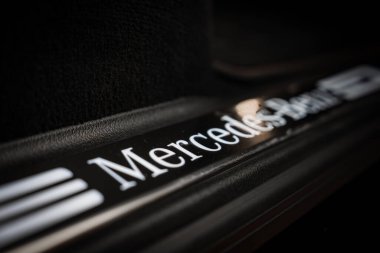 Deri kaplı siyah iç malzemelerle çevrili, parlak siyah bir yüzey üzerinde beyaz ışıklı markalı Mercedes Benz kapı pervazının yakın görüntüsü.