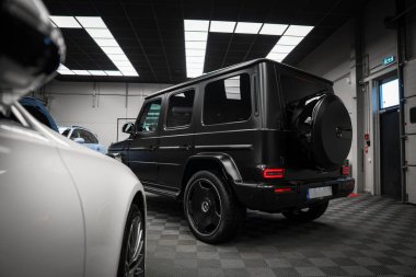 Pürüzsüz siyah Mercedes AMG sınıfı SUV. Damalı zeminde, arka ve yan profil görünüyor. Parlak ızgara desenli ışıklar altında..