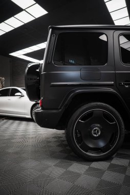 Siyah Mercedes AMG sınıfı SUV arka lastik kapağı ve alaşımlı tekerlekleri var. Arka planda beyaz lüks araba modern ışıkların altında kareli zeminde..
