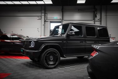 Gösterişli siyah bir Mercedes AMG sınıfı kareli zemin döşemesi, parlak ışıklandırması ve Ford Mustang GT 5.0 'ın kısmi görüntüsü olan bir sergi salonunda sergileniyor..