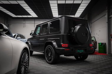 Gösterişli siyah Mercedes AMG sınıfı SUV kırmızı stop lambalı ve kareli zeminde yedek lastiği var. Beyaz araba iyi aydınlatılmış bir galeride kısmen görülebilir..