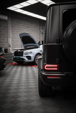 Modern bir galeride arka farları ve yedek lastik kılıfı olan siyah bir Mercedes AMG sınıfı. Arka planda kaputu açık beyaz bir Bentley var..