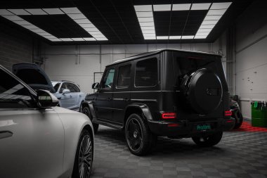 Arka planda diğer araç ve ekipmanlarla birlikte parlak tavan lambalarının altında kareli bir zemine park etmiş siyah bir Mercedes AMG sınıfı SUV..