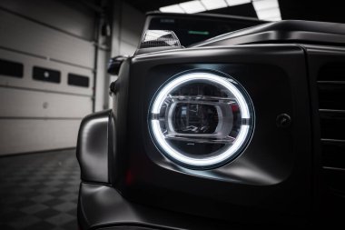 Dairesel LED ışık, mat siyah kaplama ve kapalı sergi salonu yansımaları ile Mercedes AMG sınıf far yakın görünüm.