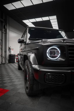 Siyah Mercedes AMG sınıfı SUV, mat kaplama, dairesel LED far, ve modern bir sergi salonunda kalın ızgara desenli siyah zemin.