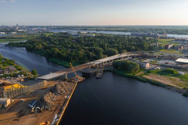 Kısmen tamamlanmış bir köprü Riga, Letonya 'da bir nehri vinçler, inşaat malzemeleri ve bir tersaneyle kaplar. Şehir manzarası ufukta görünüyor..