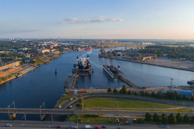 Riga, Letonya 'da bir tersane, rıhtımlı bir gemi, vinçler ve endüstriyel yapılar. Daugava Nehri, gün batımını yansıtır. Köprüler ve şehir manzarası görünür..