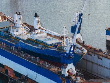 Mavi beyaz bir kargo gemisi Riga tersanesine yanaştı. Etrafı vinç ve ekipmanlarla çevrili. Sakin su endüstriyel deniz ortamını yansıtır..